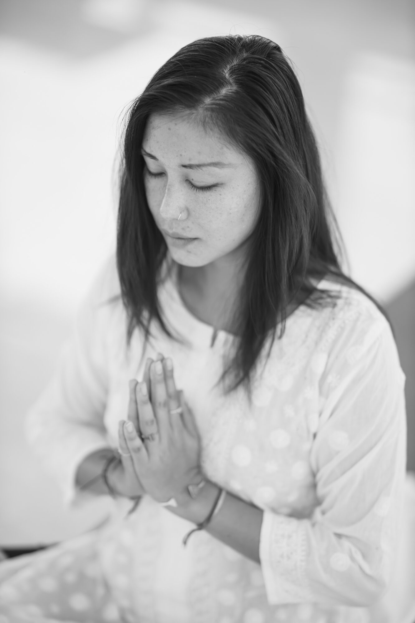 Young woman with closed eyes and hands pressed together in prayer or meditation, wearing a light-colored dress.