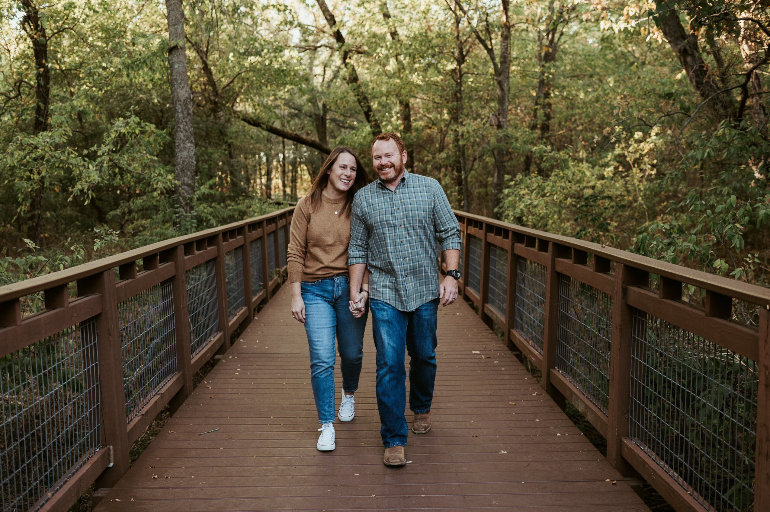 A smiling couple walks arm-in-arm on a wooden bridge surrounded by trees in a forest.