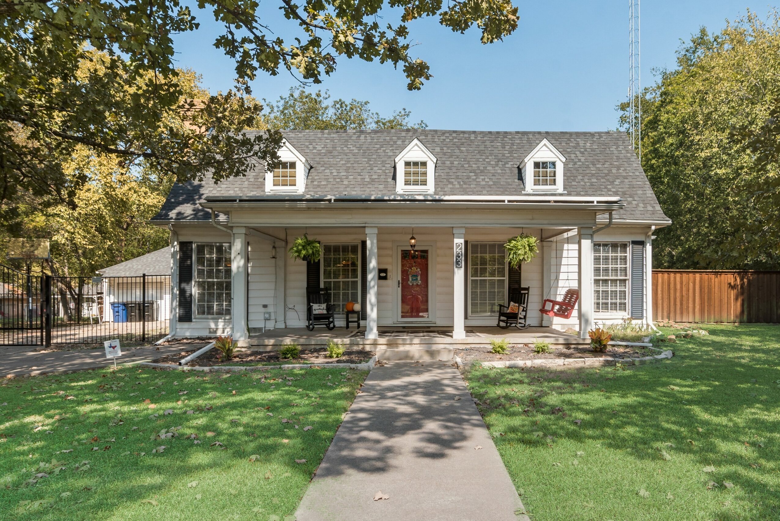 Front view of a house with a porch, two chairs, and a walkway leading to the door, surrounded by trees and grass.