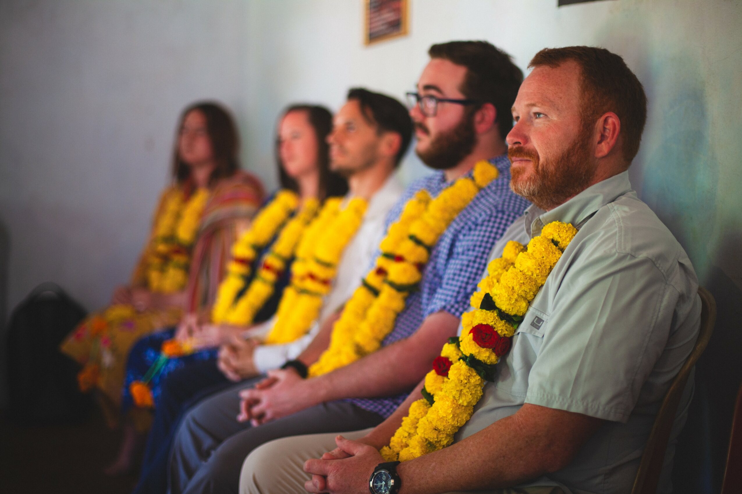 Five people sit in a row, wearing yellow flower garlands, against a light-colored wall.