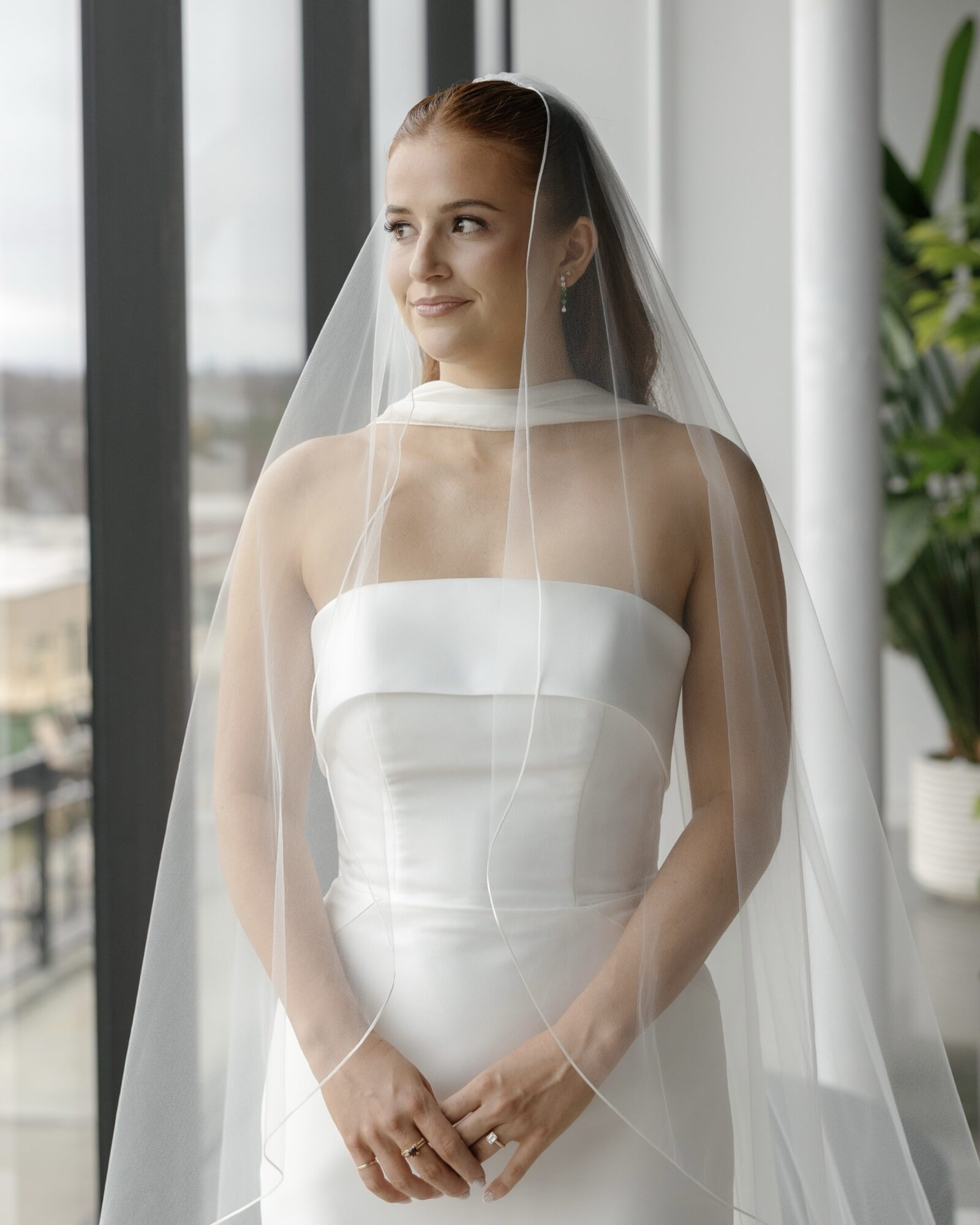 Bride in a strapless white wedding dress with veil, standing indoors near large windows, smiling softly, hands clasped.