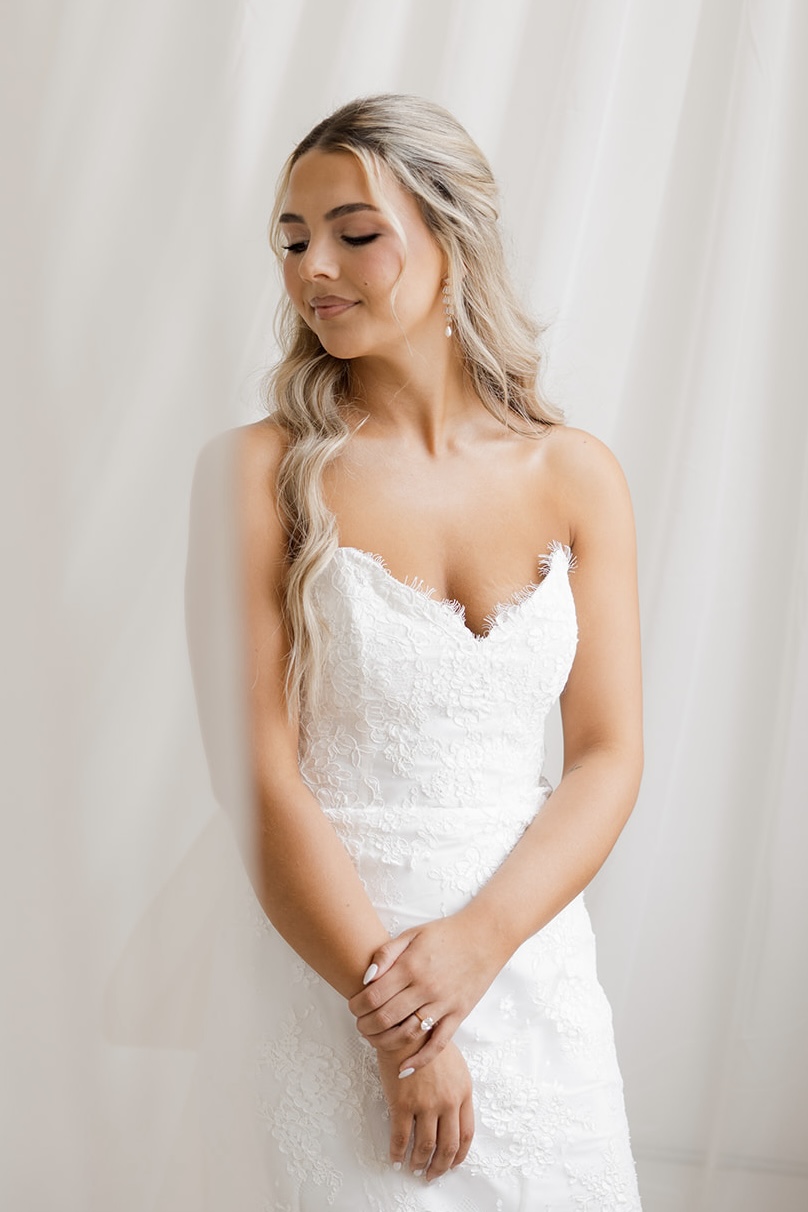 Young woman in a white lace dress with long, wavy blonde hair, standing with eyes closed and hands clasped.