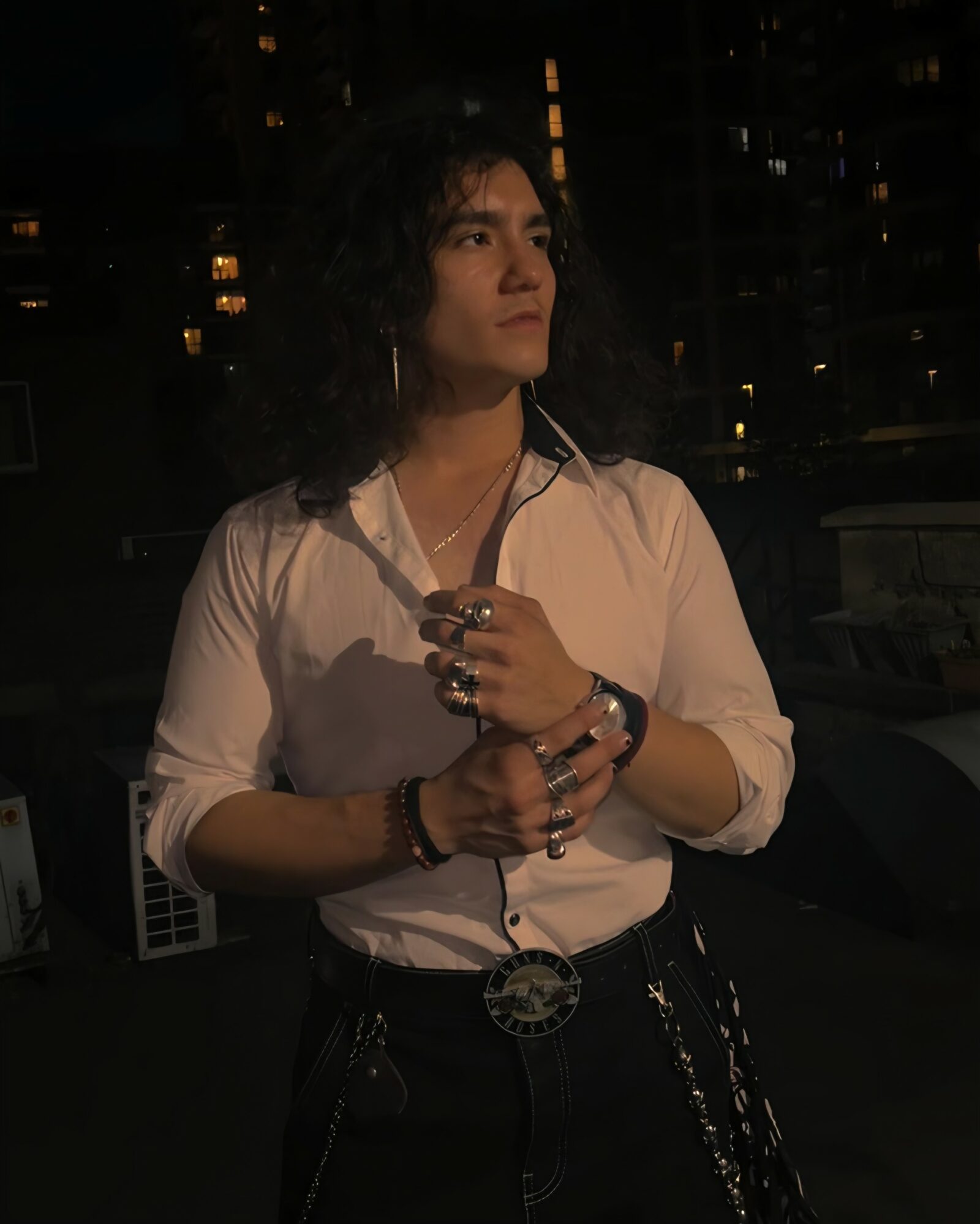 Woman with curly hair wearing a white shirt, standing outdoors at night with city lights in background.
