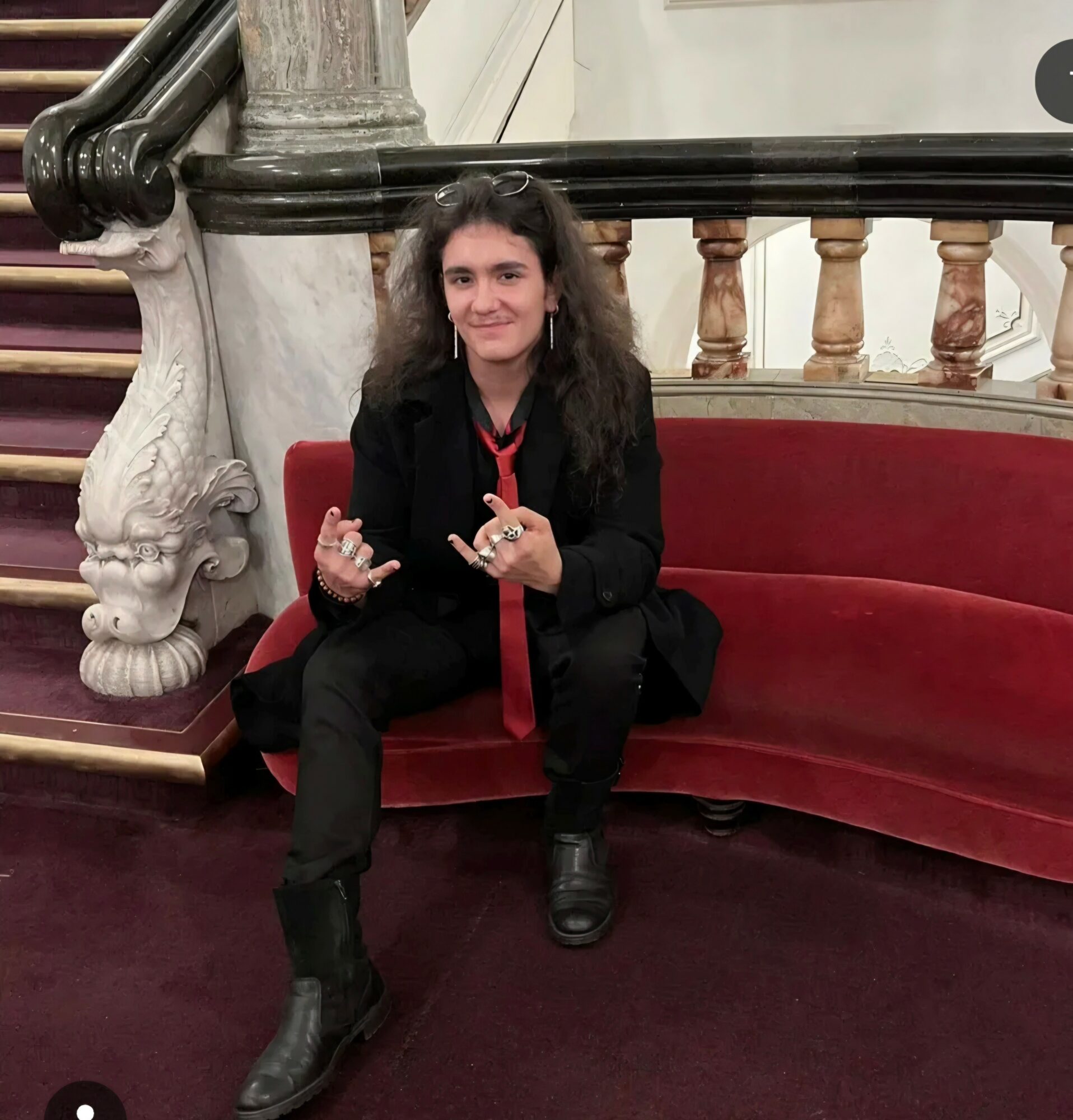Woman with curly hair sitting on a red curved sofa, smiling, in an ornate interior with marble and wood details.