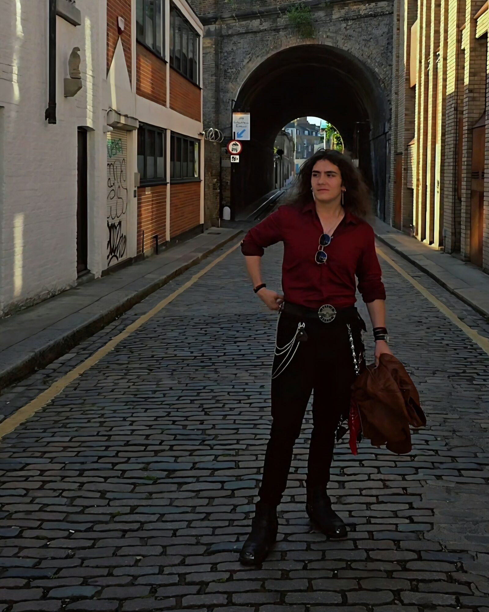 Woman standing on cobblestone street under archway, wearing red shirt, black pants, sunglasses, holding brown jacket.