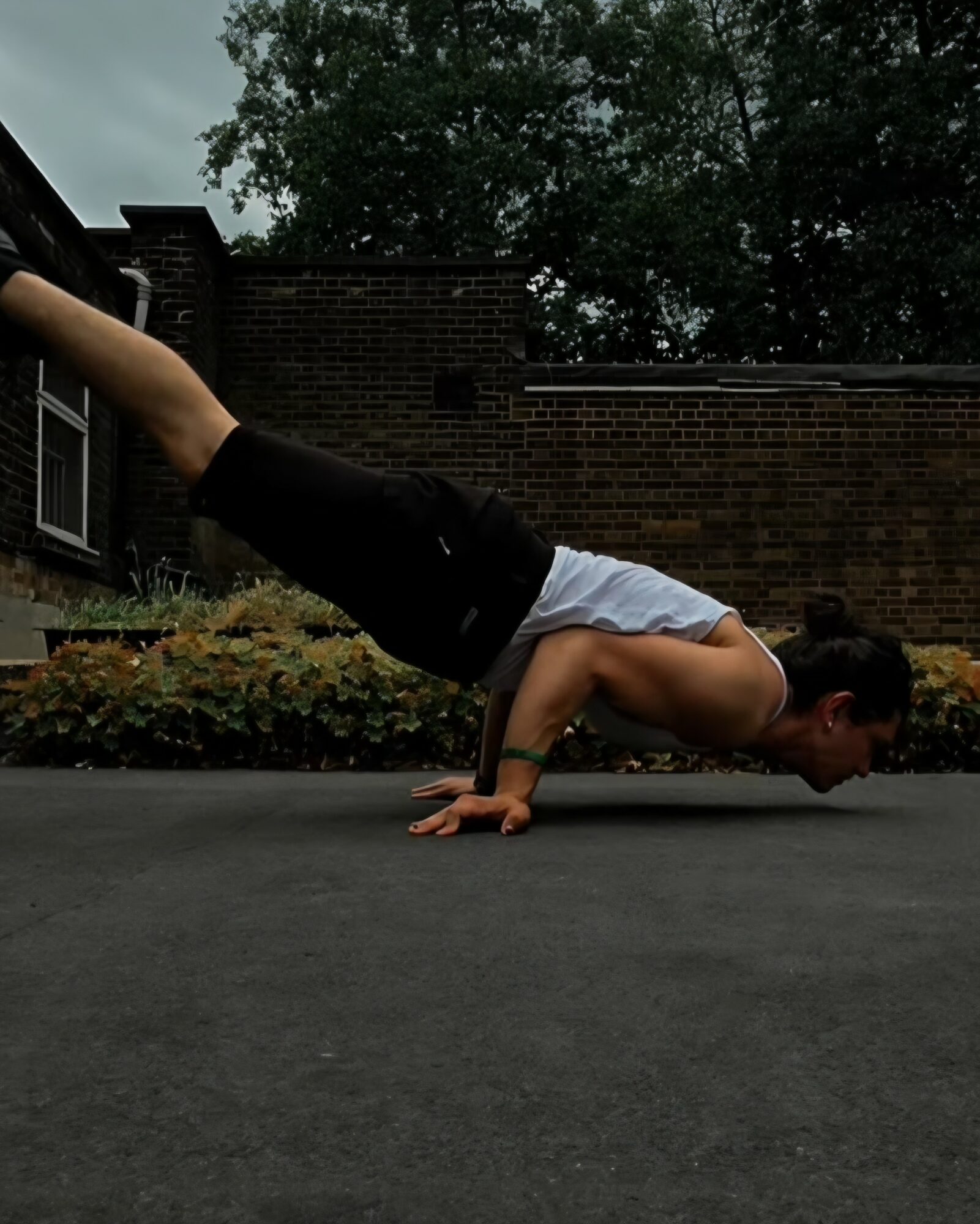 Person performing a push-up outdoors on a paved surface with a brick building and trees in the background.