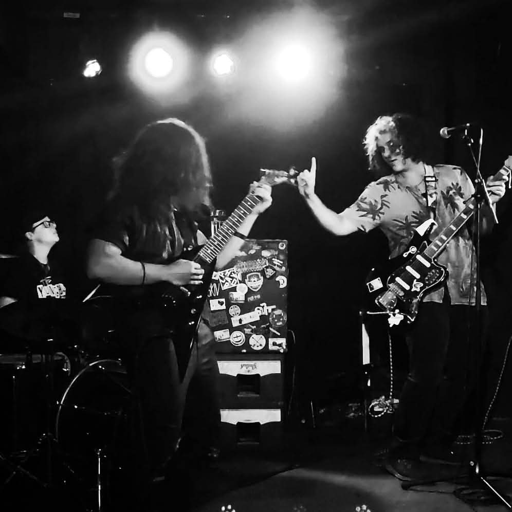 Three musicians perform on stage with guitars, one reaching out to another, illuminated by stage lights, in black and white.