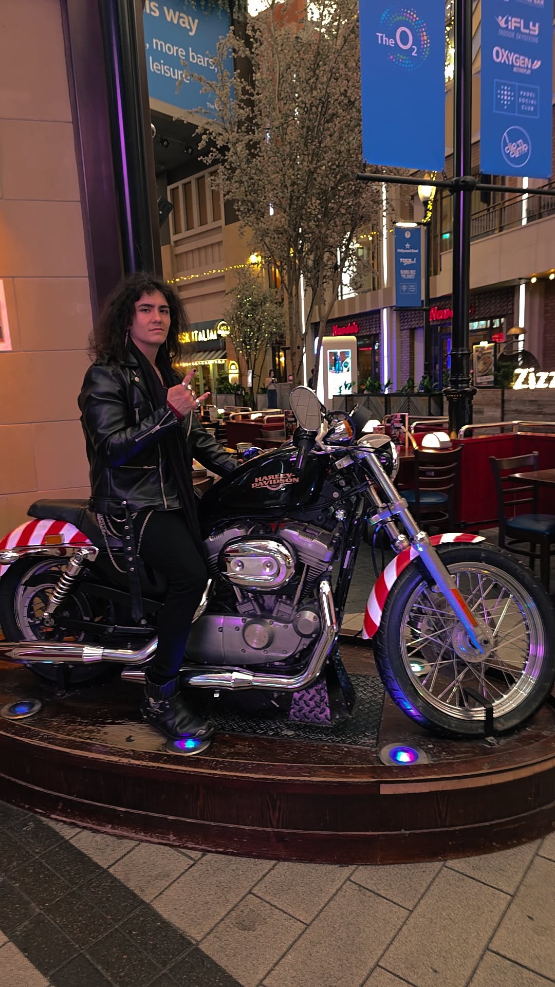 Person sitting on a motorcycle in an outdoor shopping area at night, with illuminated signs and trees in the background.