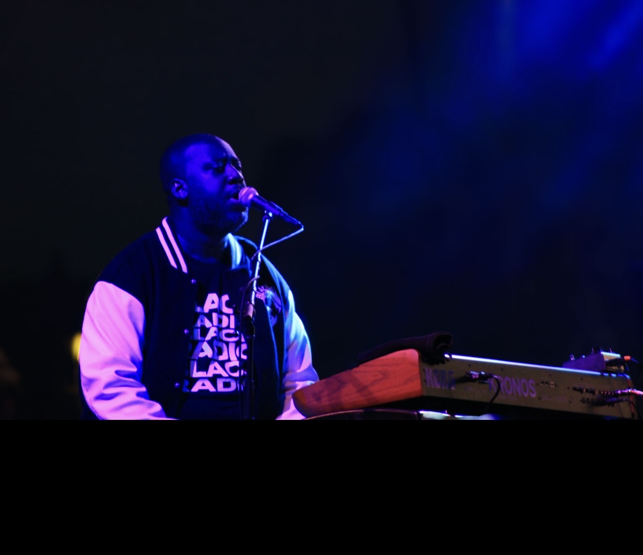 Person singing into a microphone with a keyboard in front, illuminated by blue stage lights.