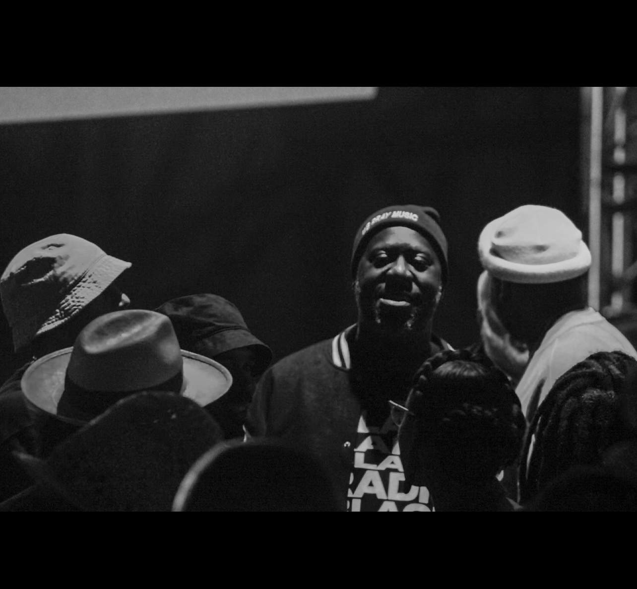Group of people gathered, with one person speaking or performing, in black and white. The scene appears indoors.