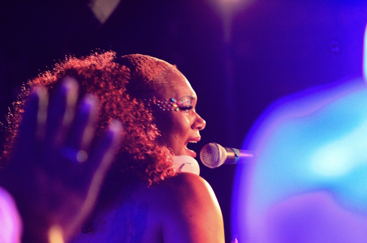 Person singing into a microphone with colorful stage lighting, curly hair, and a hand raised nearby.