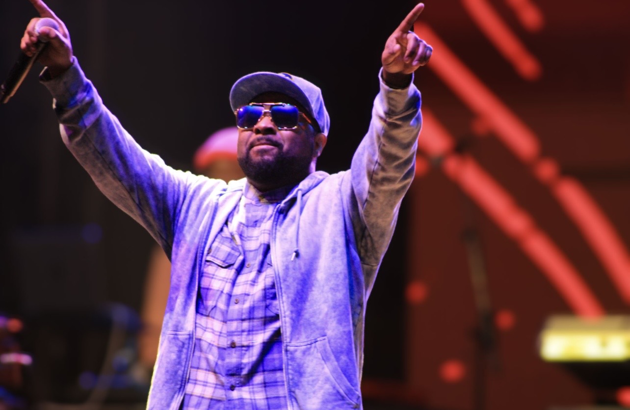Person wearing sunglasses and a cap, raising both arms on stage with red lights in background.