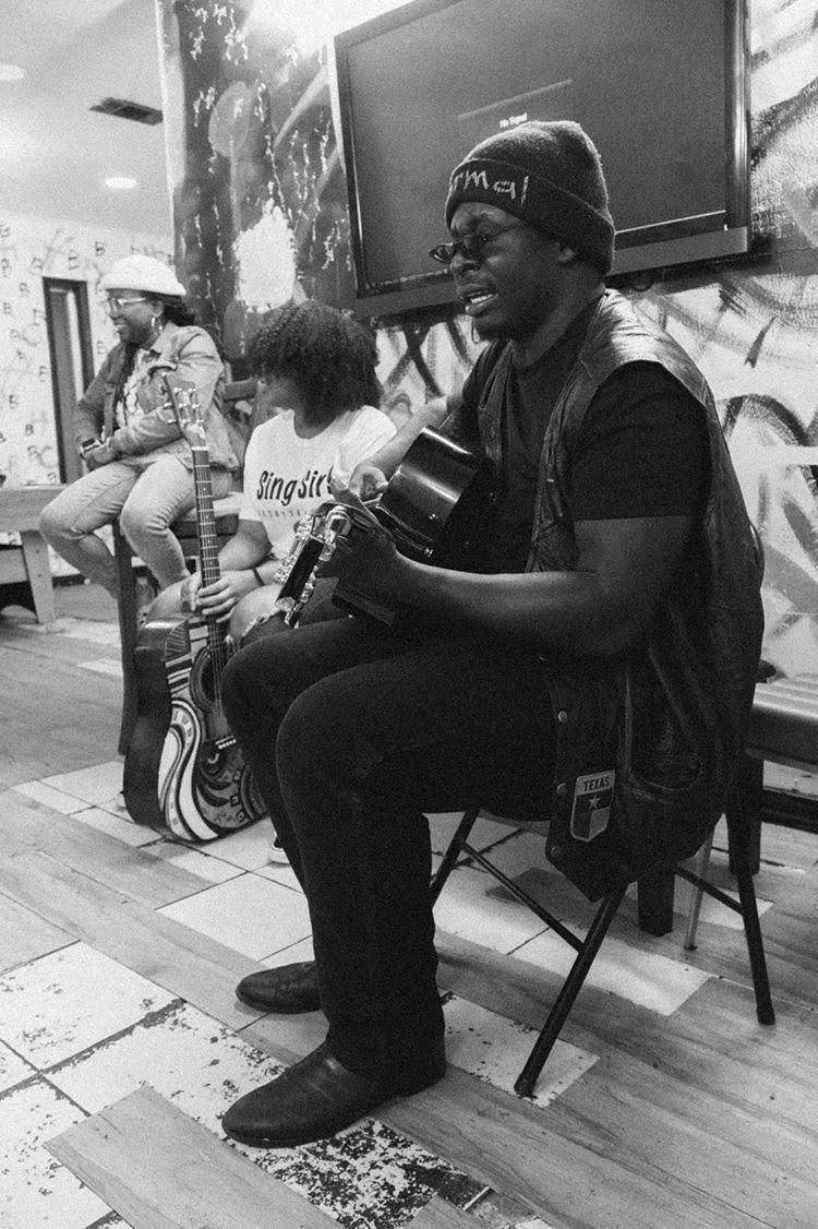 Person playing guitar in a room with others sitting nearby, some wearing hats, and a large screen on the wall.