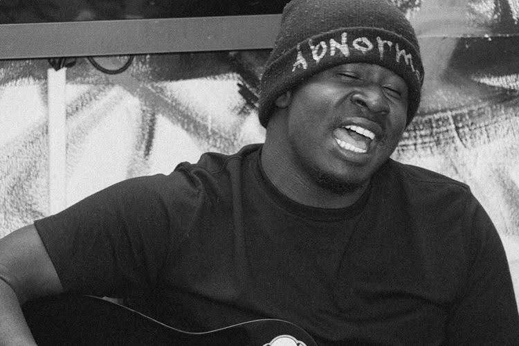 Person wearing a beanie and black shirt, making a facial expression, holding a guitar, in black and white.