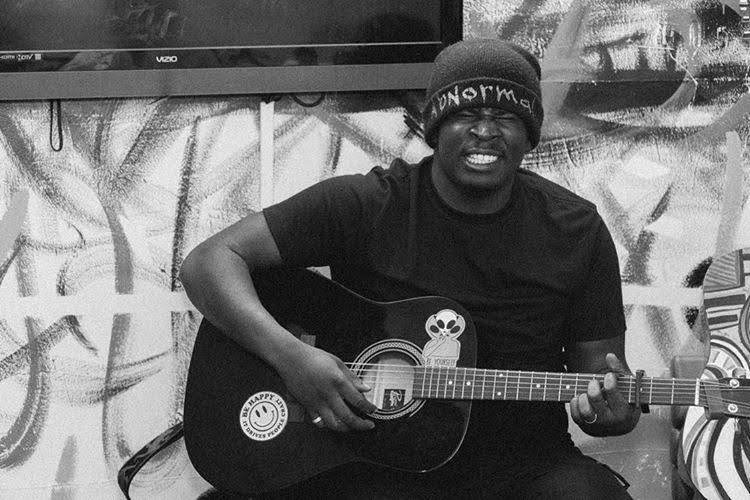 Person wearing a beanie playing an acoustic guitar, smiling, with graffiti background, in black and white.