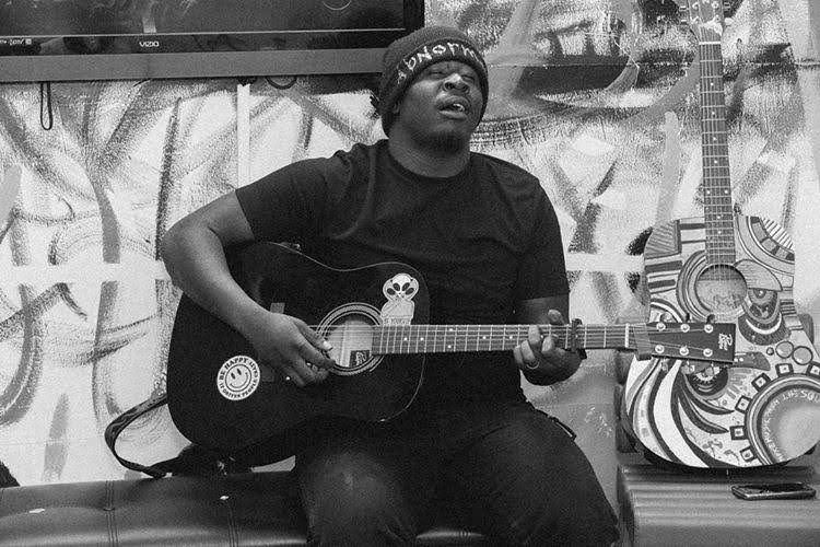 Person playing guitar in a room with graffiti and decorated guitars on the wall, wearing a headband and glasses.