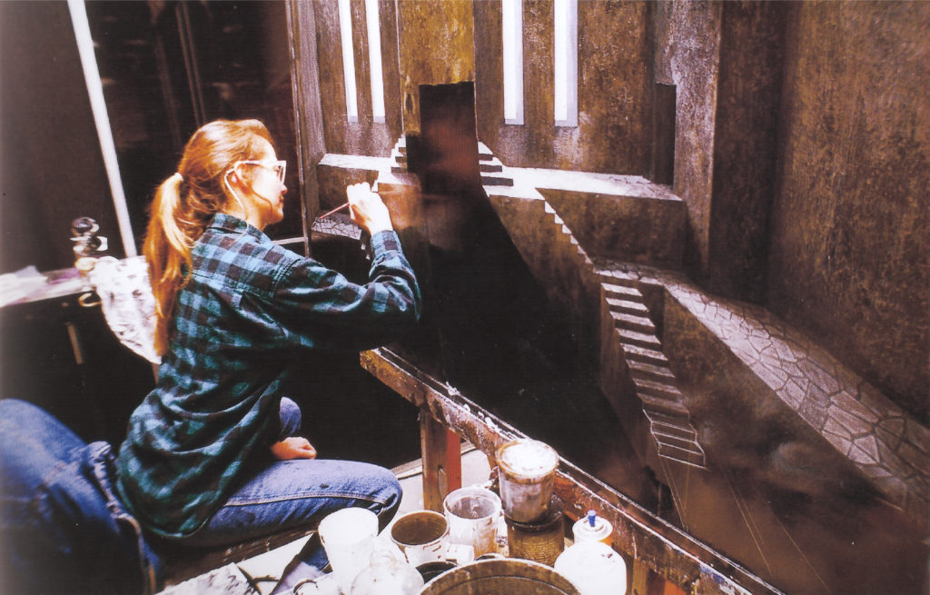 Person with long hair and plaid shirt sitting at a table, taking a photo inside a rustic wooden structure.