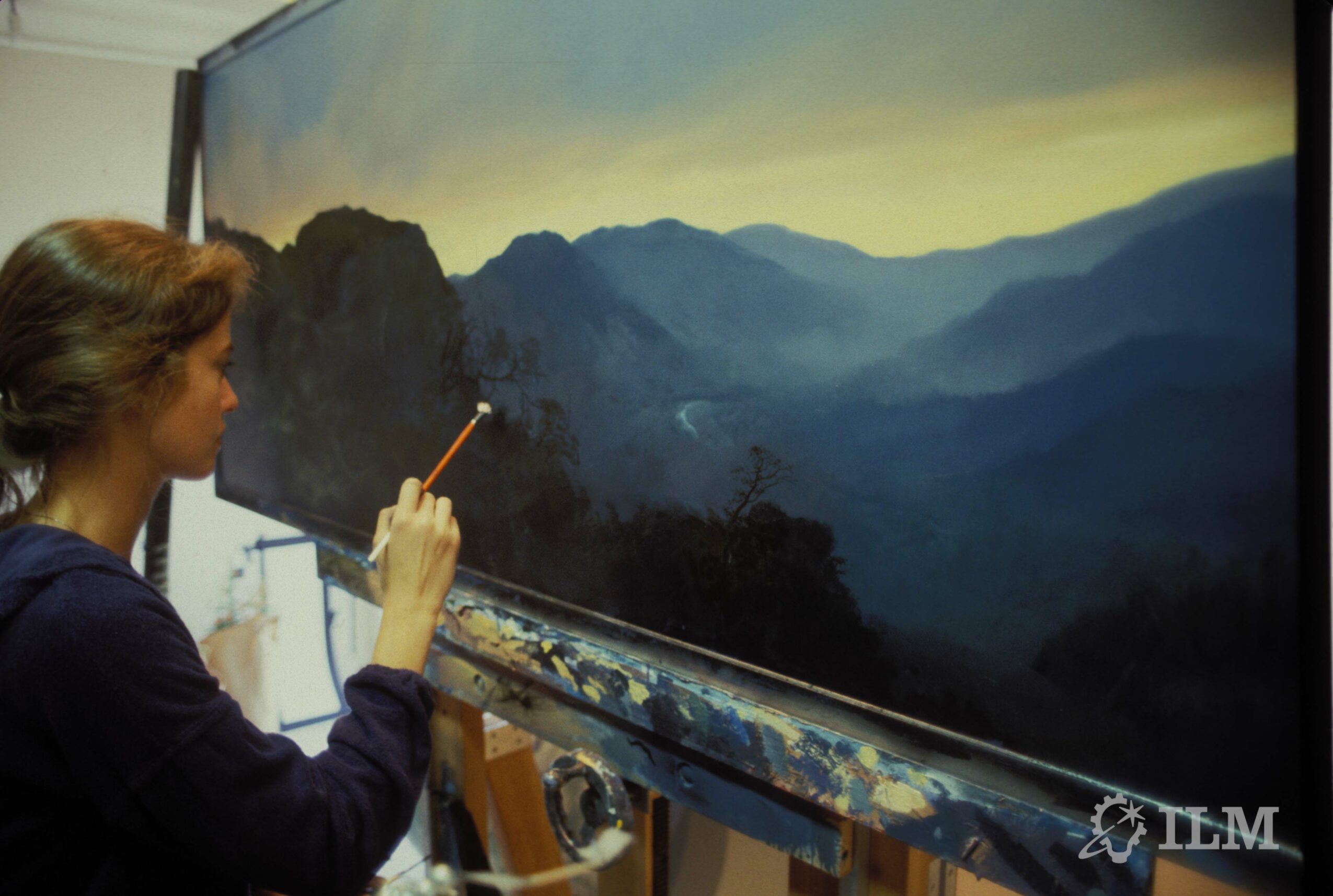 Person painting a landscape on a large canvas with mountains and sky, using a brush, in an indoor studio.