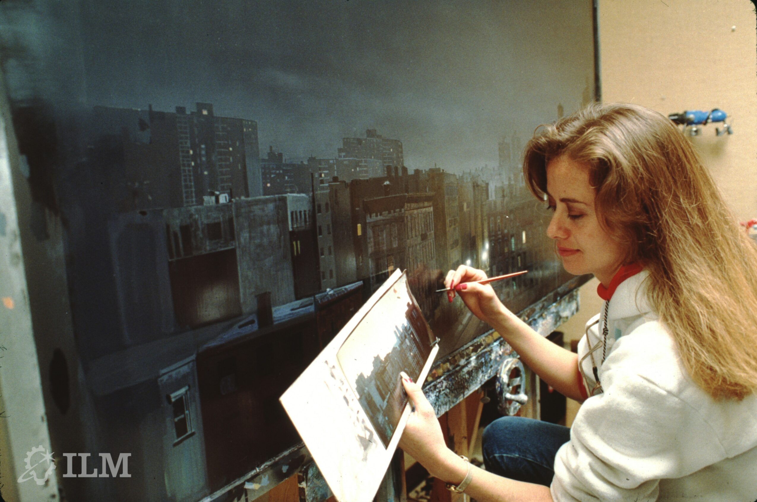 Woman painting on a large blackboard with cityscape image, holding a palette and brush, in a room with beige wall.