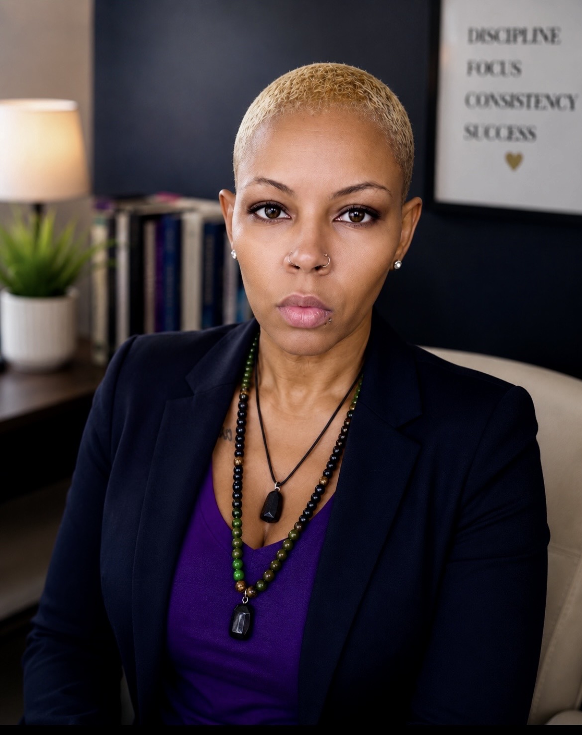 Woman with short blonde hair wearing a black blazer and purple top, sitting in an office with a motivational poster behind her.