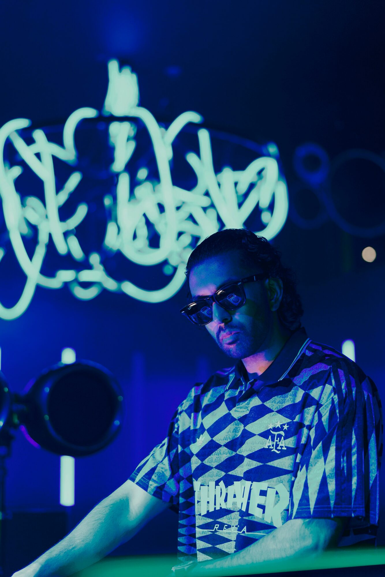 Person wearing sunglasses and a patterned shirt, standing in front of a blue graffiti-style background, with a dark setting.