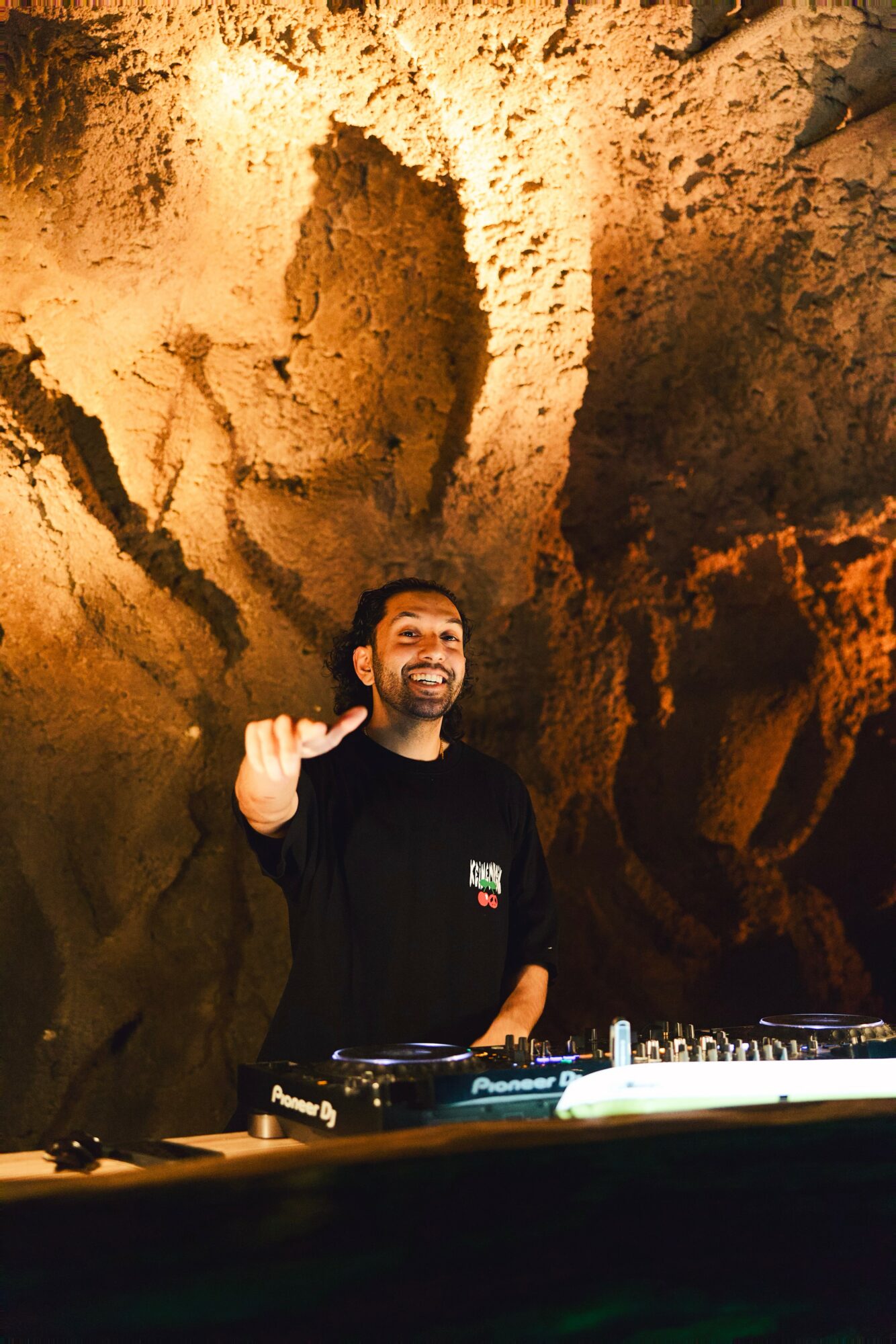 Person standing in a cave with rocky walls, smiling and waving, with DJ equipment in front.