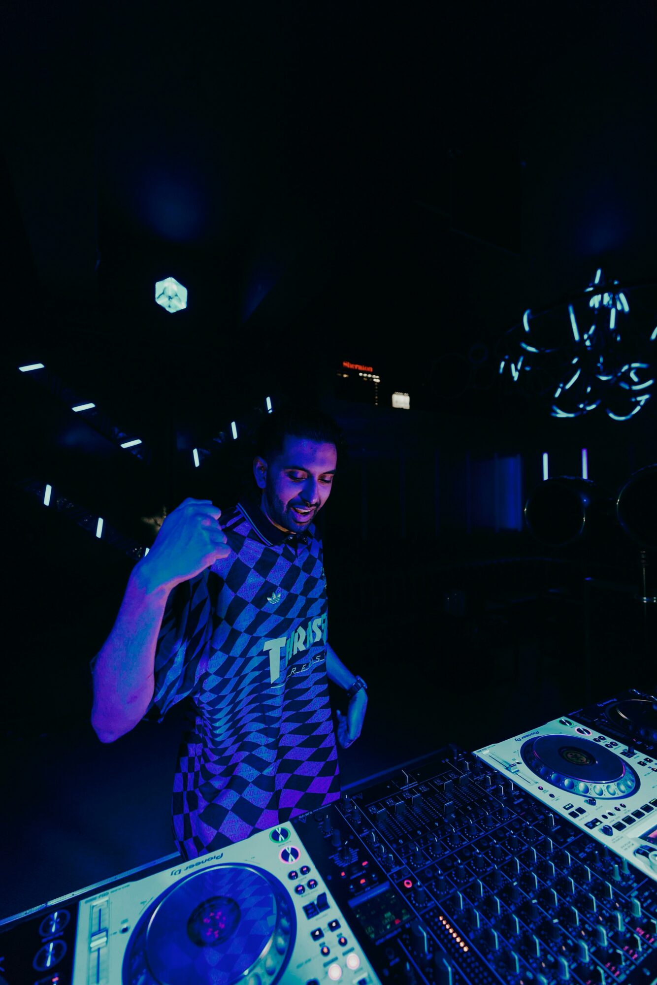 Person with a beard smiling and raising a fist in a dark, illuminated environment with DJ equipment nearby.