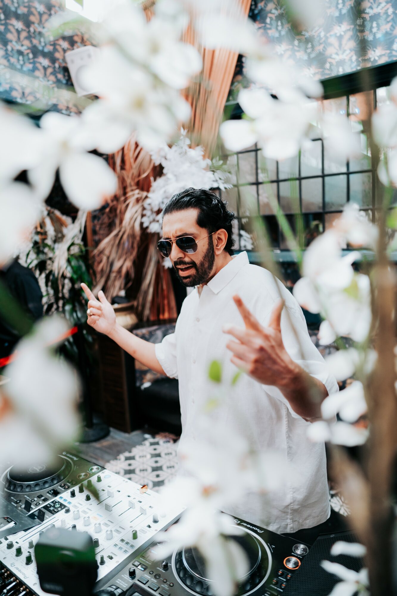Man with sunglasses and beard smiling and making a peace sign, surrounded by white flowers and greenery.
