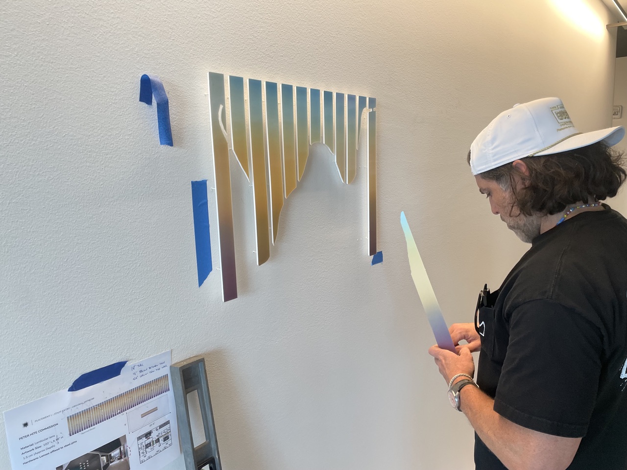 Person with a white cap and black shirt looking at a colorful wall art piece, holding a smartphone, in a well-lit room.