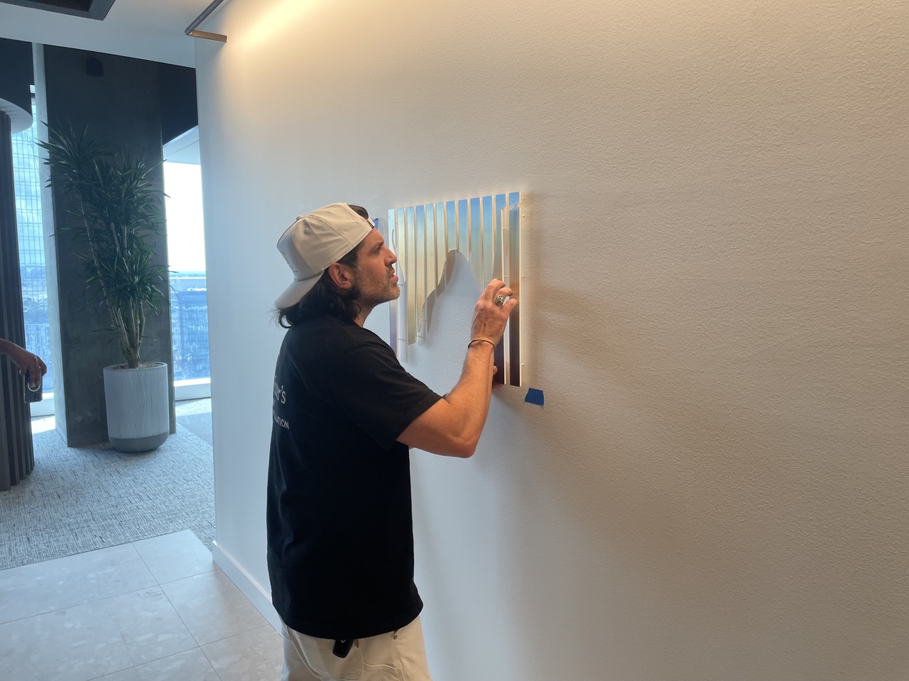 Person with a cap painting on a wall indoors, with a window and outdoor view in the background.