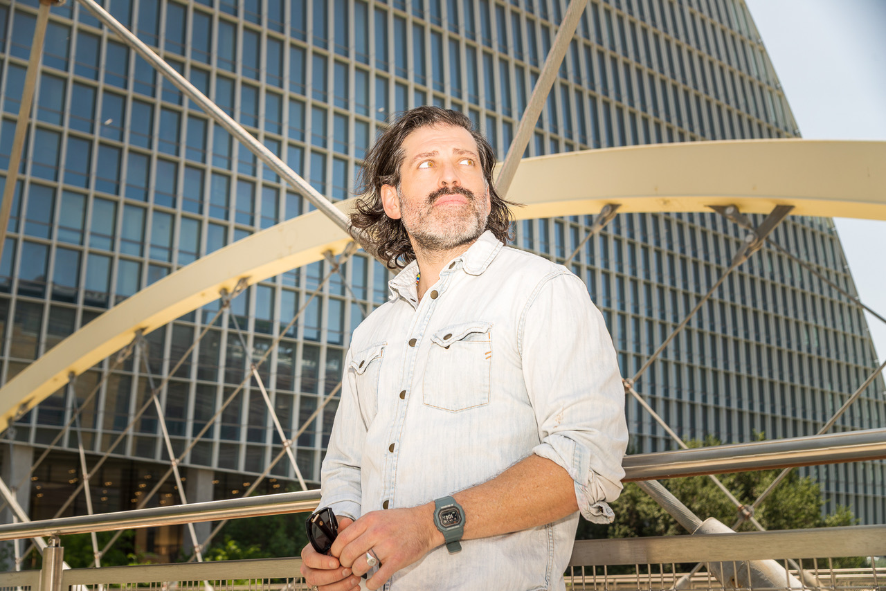 Man with long hair and beard wearing a white shirt standing outdoors in front of modern glass buildings and a large yellow structure.