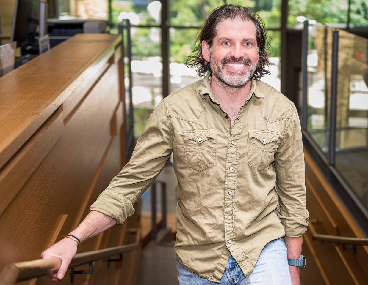 Man with curly hair and beard smiling, standing indoors near a wooden railing, with large windows and greenery outside.