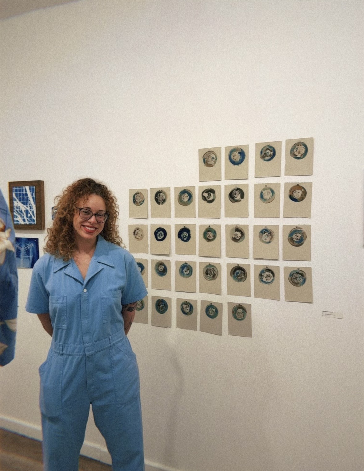 Woman with curly hair in blue jumpsuit standing in front of wall with framed artwork and circular objects, smiling.