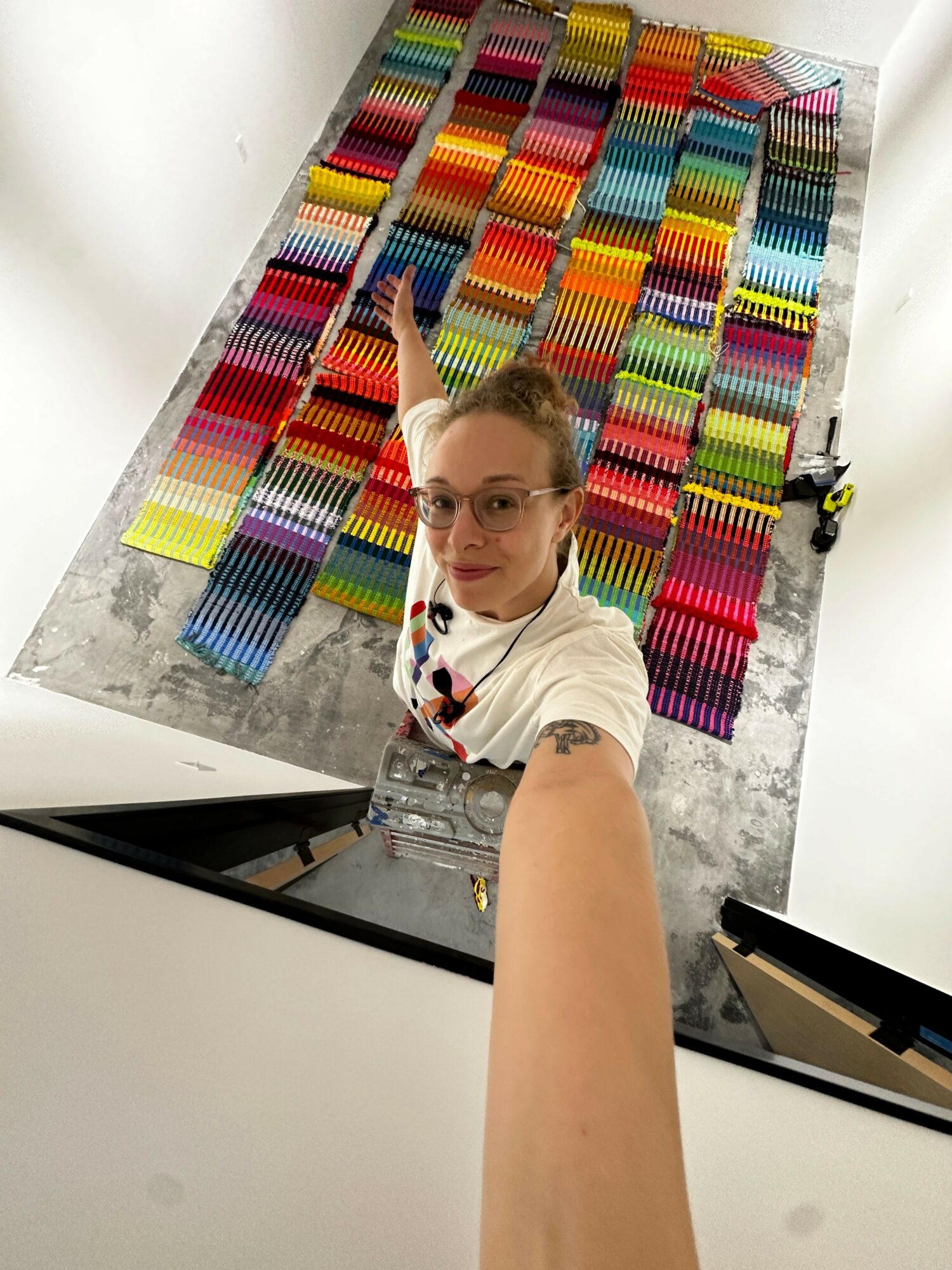 Person taking a selfie in front of a colorful wall display of stacked, multicolored objects, with a white shirt and a tattoo on the arm.