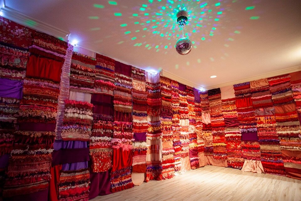 Room with walls covered in multicolored fabric strips, ceiling with disco ball and colorful light patterns, wooden floor.
