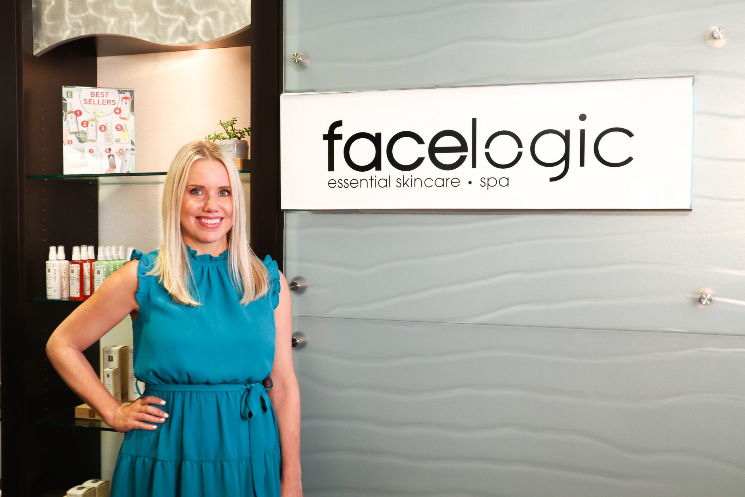 Woman with blonde hair in a blue dress standing next to a sign that says 'facelogic' in a spa or skincare clinic.