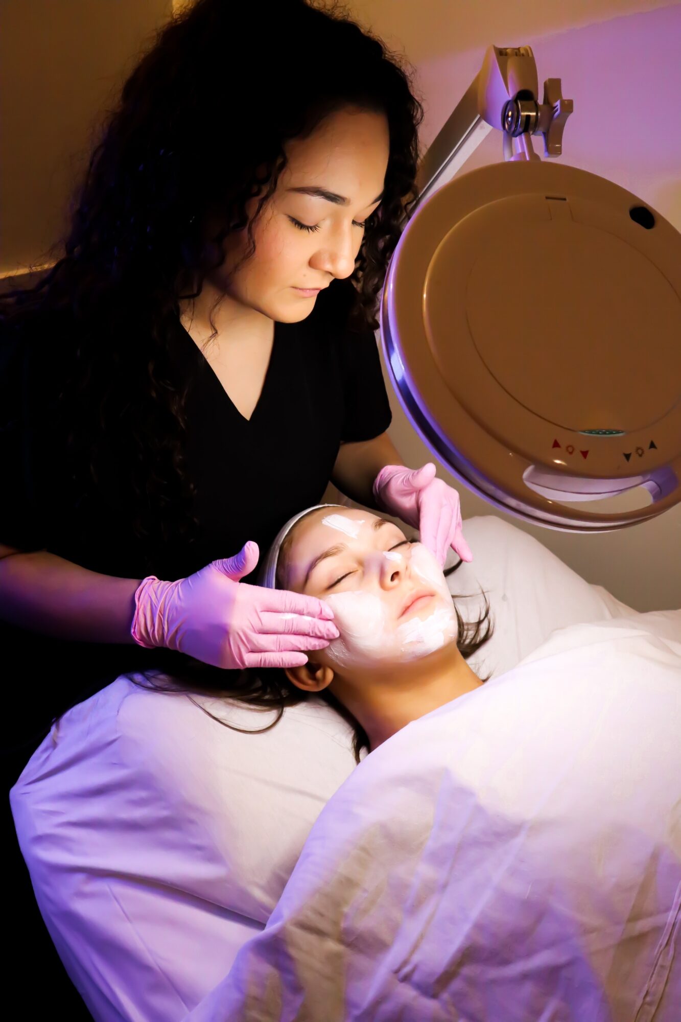 A woman wearing pink gloves performs a facial treatment on a woman lying with eyes closed, in a clinical setting.