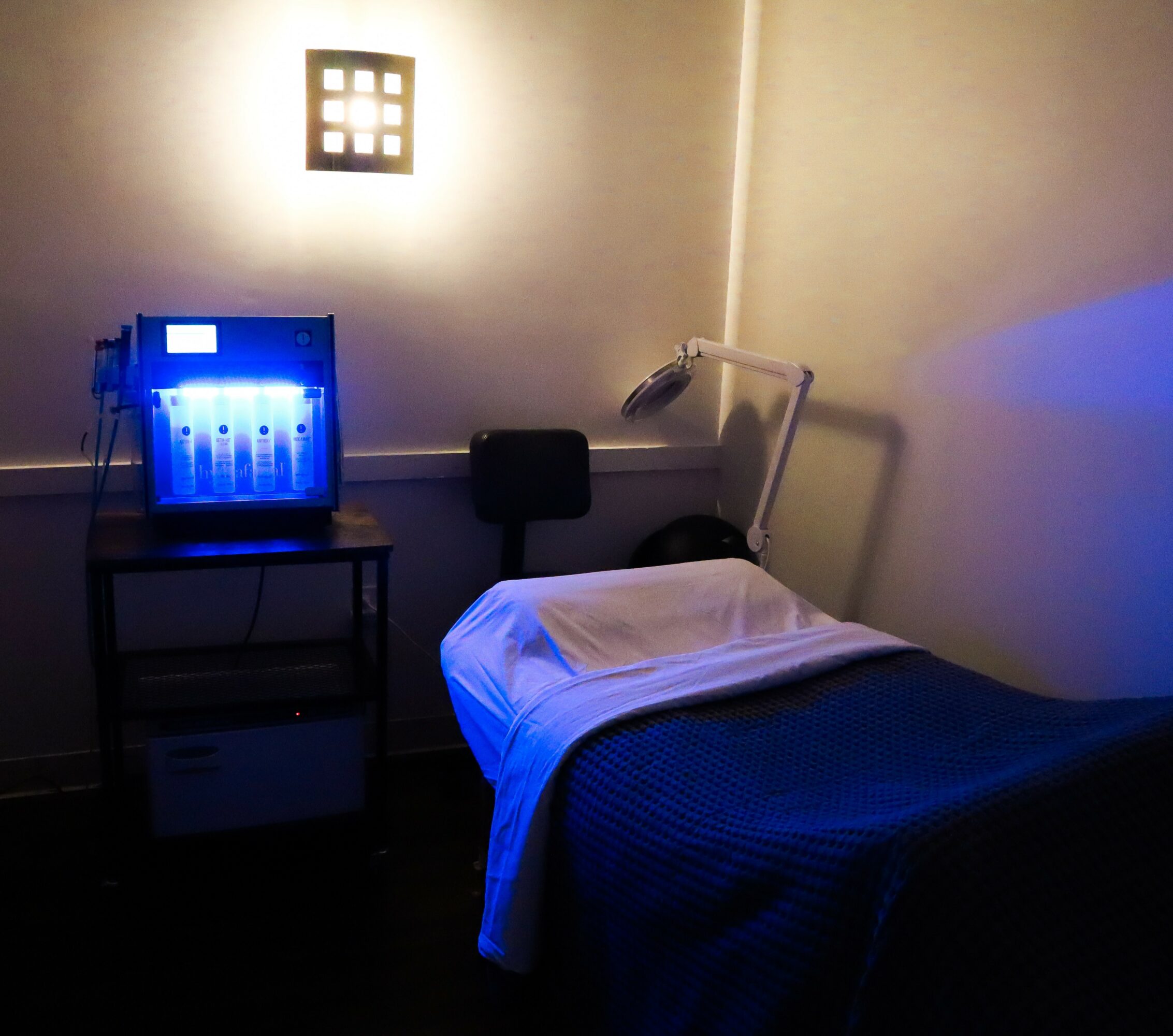Hospital bed with white pillow and blanket, medical equipment on a table, dim lighting, wall light fixture, and a lamp.