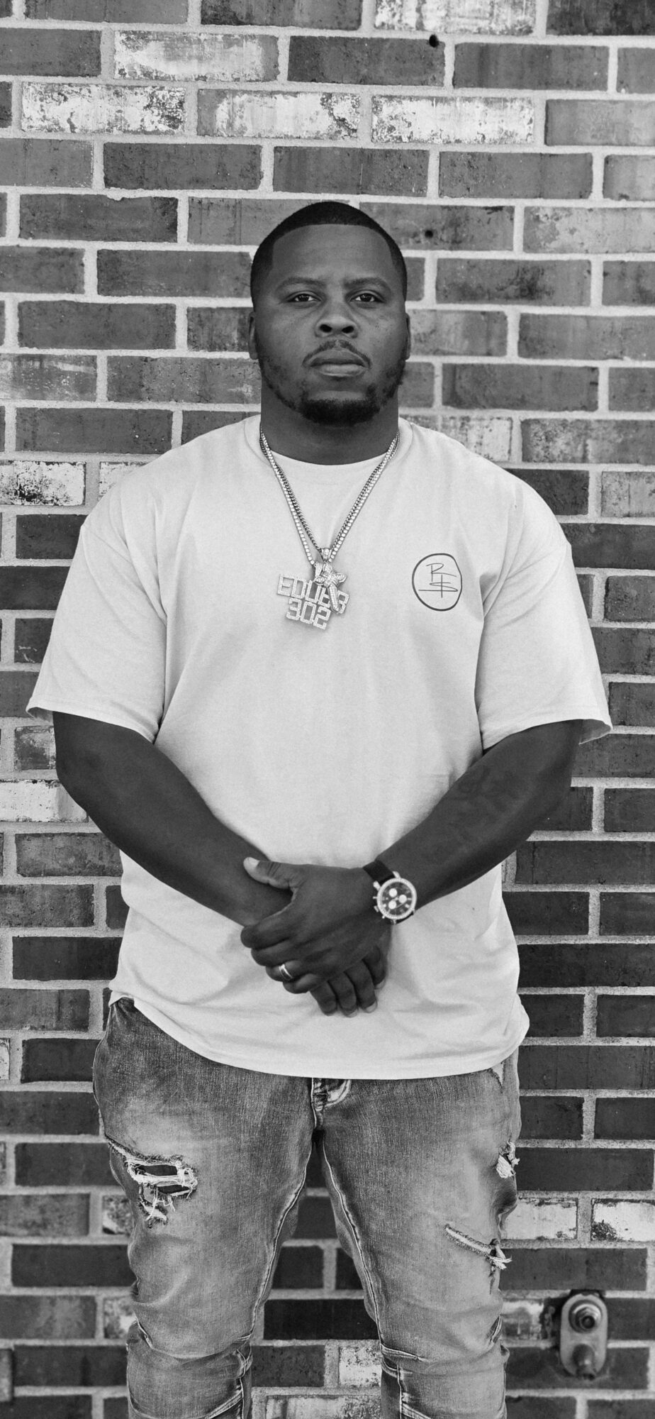 Man with short hair and beard standing against a brick wall, wearing a t-shirt, ripped jeans, and jewelry.