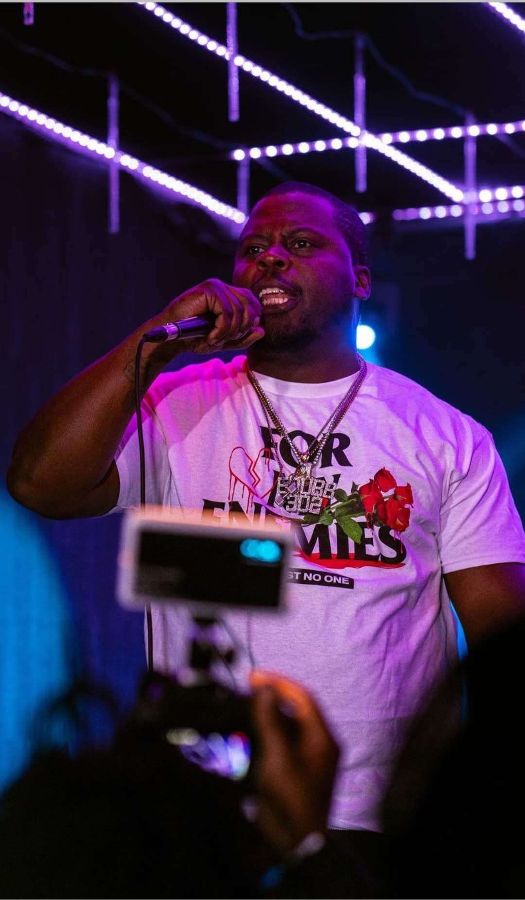 Person speaking into microphone on stage with purple lighting and ceiling lights, wearing a white T-shirt with text and graphics.