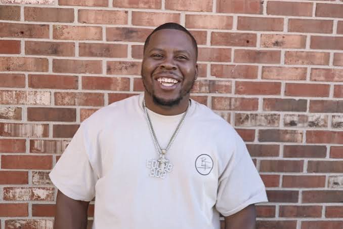 Smiling man with short hair and a beard, wearing a light-colored T-shirt and a necklace, standing in front of a brick wall.