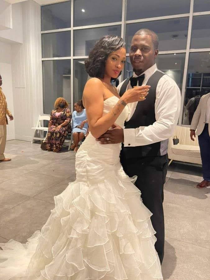 A woman in a white wedding dress and a man in a black vest and white shirt pose together indoors, with other people in the background.