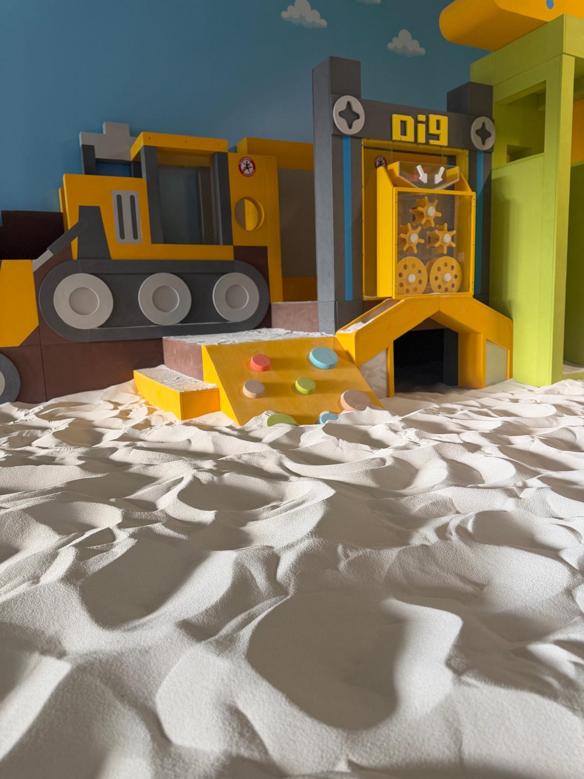 Colorful toy construction site with a slide, truck, and sand area, set against a blue wall background.