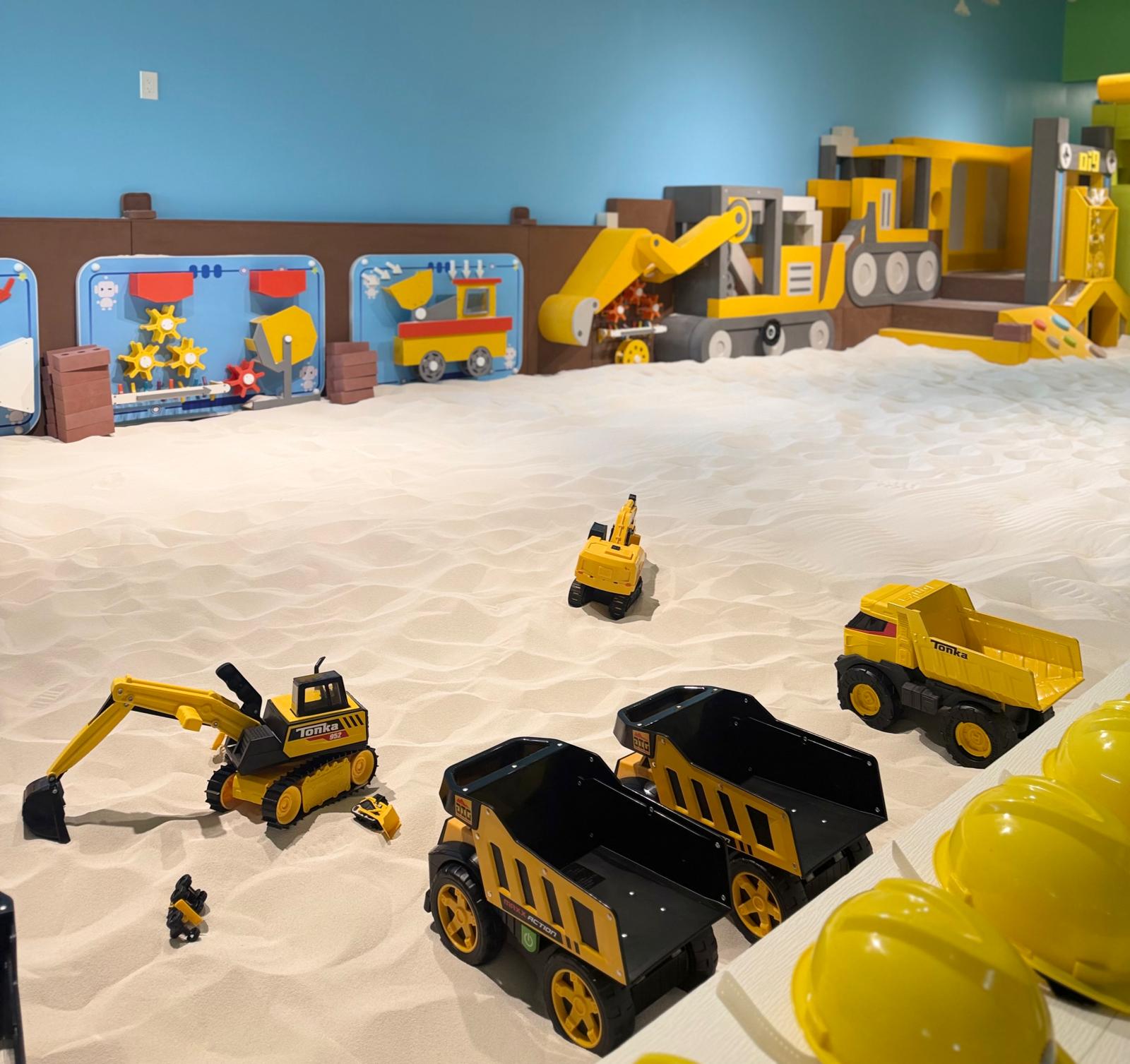 Toy construction vehicles on sand with a blue wall and colorful train in background.