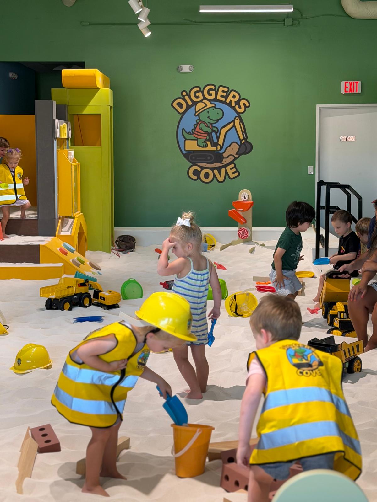 Children playing in an indoor sandbox with toys, wearing yellow and blue striped vests and yellow hats, in a colorful play area.