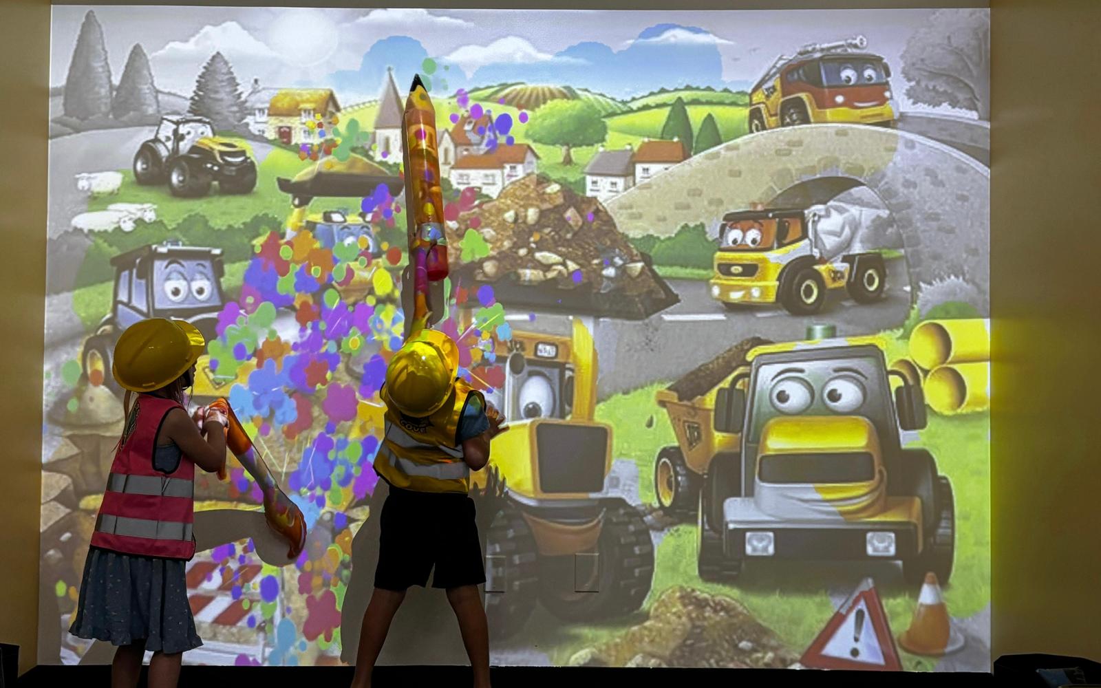 Two children interact with a colorful animated vehicle scene on a wall, featuring various cartoon trucks and construction vehicles.