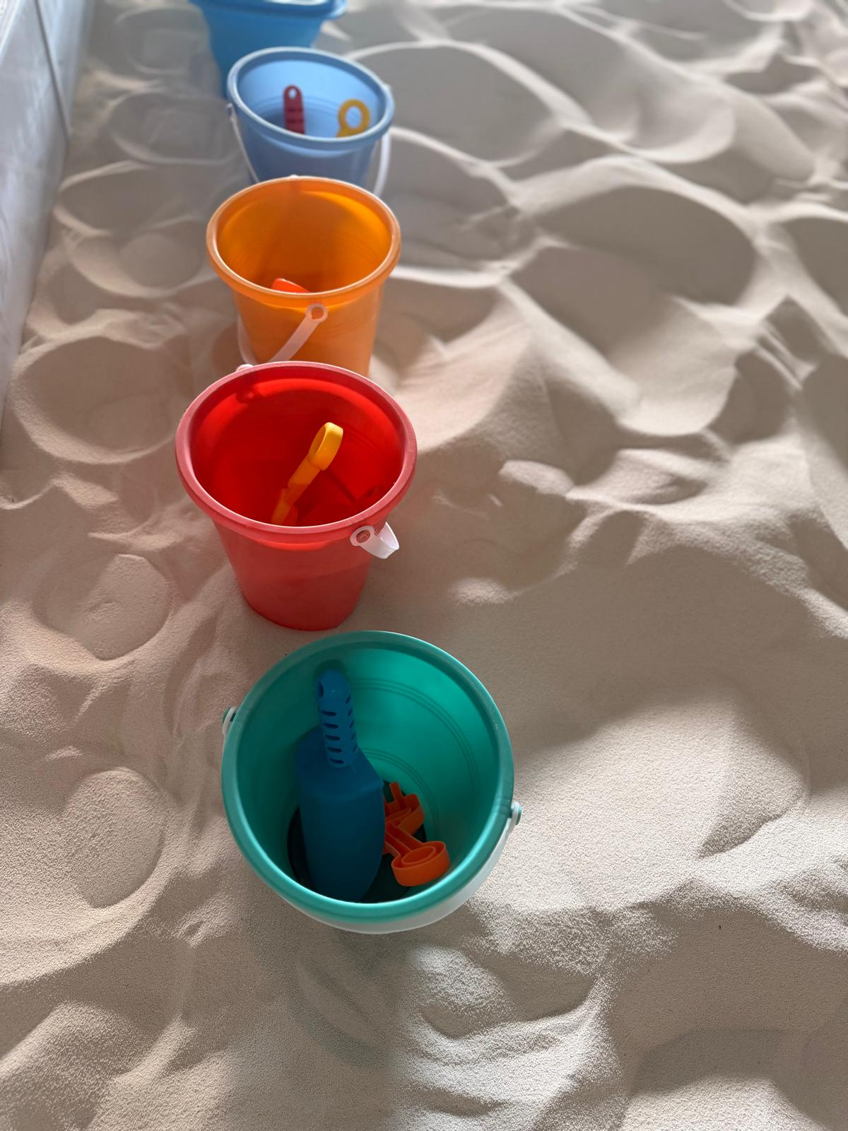 Five colorful buckets arranged in a line on sand, each with a toy inside, from blue to red to orange to teal.