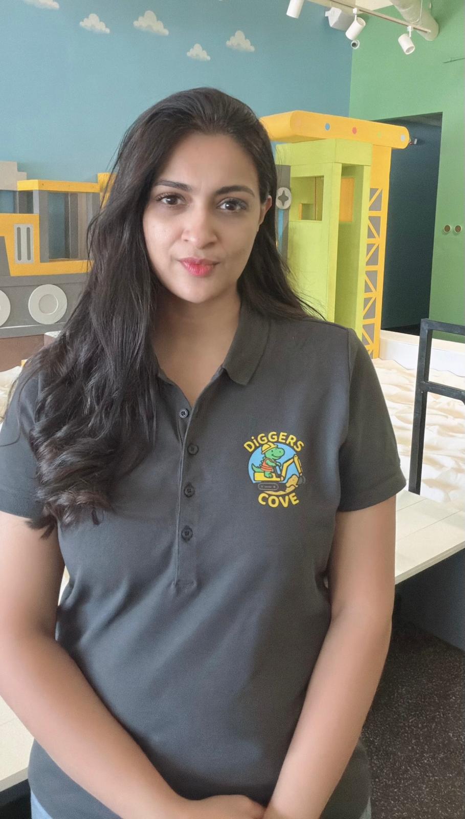 Young woman with long dark hair wearing a gray polo shirt with a logo, standing indoors.