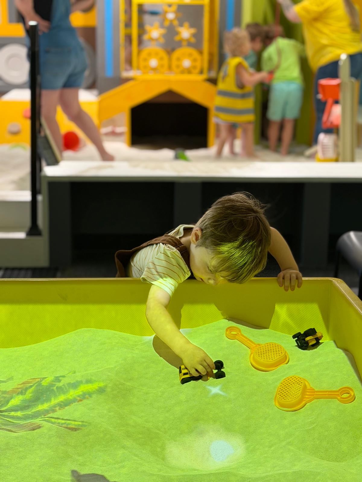 Child playing with toy guitars on a green surface, with a colorful indoor play area in the background.