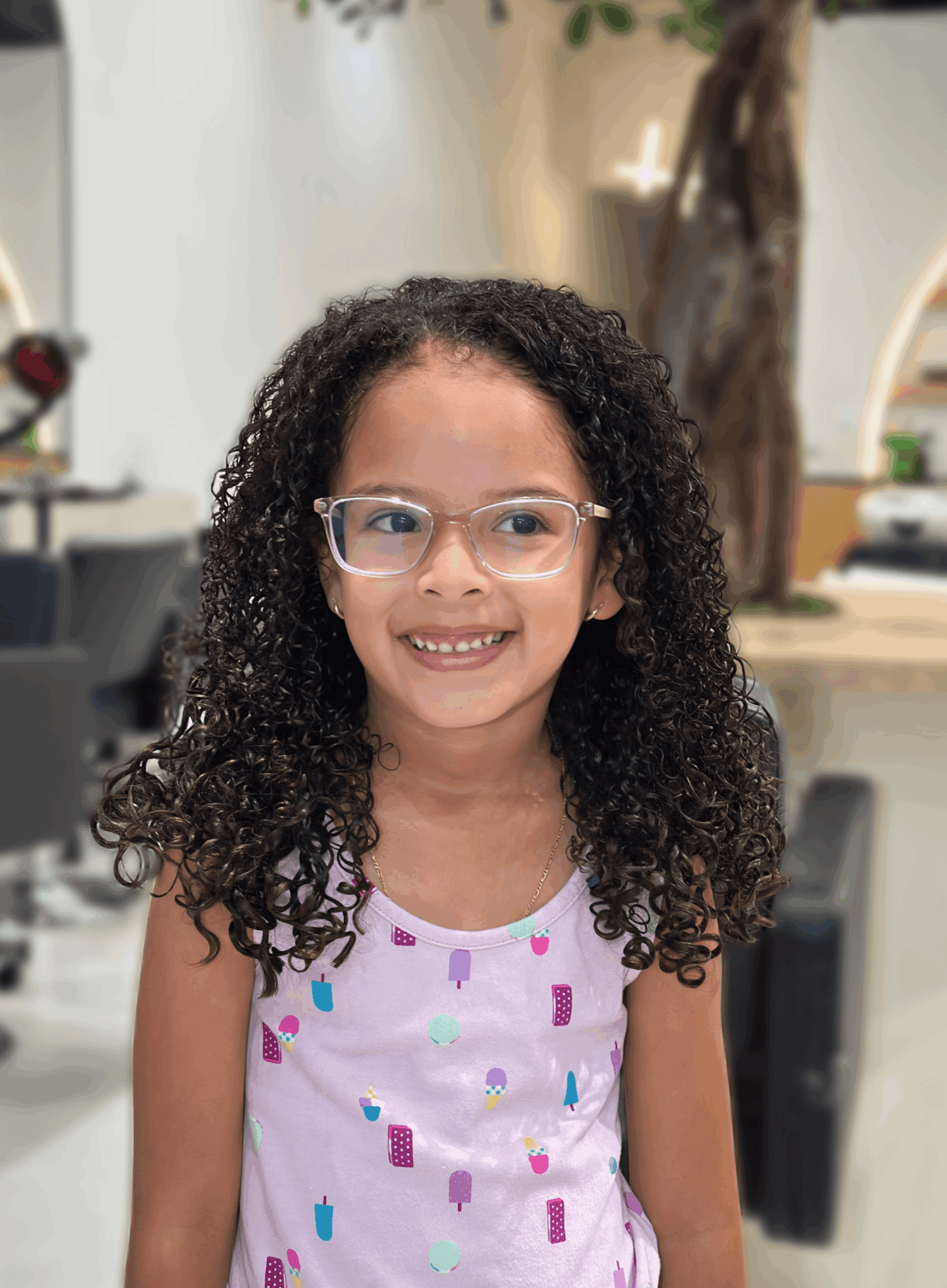 Young girl with curly hair and glasses smiling indoors, wearing a white shirt with colorful patterns.
